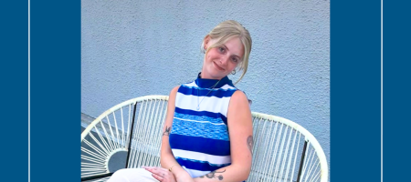 A young blond woman sits on a white wicker bench in front of a gray stucco wall. She is wearing a blue and white sleeveless blouse and and white pants. Her arms are folded in her lap and her hair is loosely pulled back. She is smiling and her head is cocked to the side.