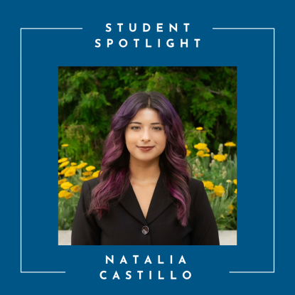 A young woman wearing a dark outfit. She has dark eyes and is smiling. Her hair is dark with a slight purple hue. In the background is greenery and yellow flowers.