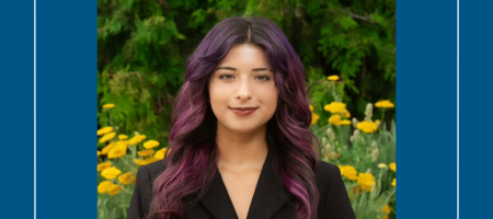 A young woman wearing a dark outfit. She has dark eyes and is smiling. Her hair is dark with a slight purple hue. In the background is greenery and yellow flowers.