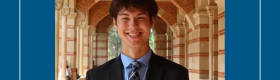 Young man in a dark suit, smiling, under the Royce Hall arches